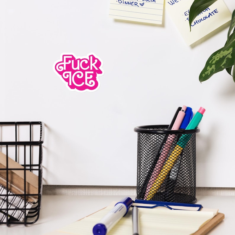 May include: A pink sticker with the words "Fuck Ice" in a stylized font. The sticker is on a white surface, with a black pen holder, pens, and a notepad in the background.