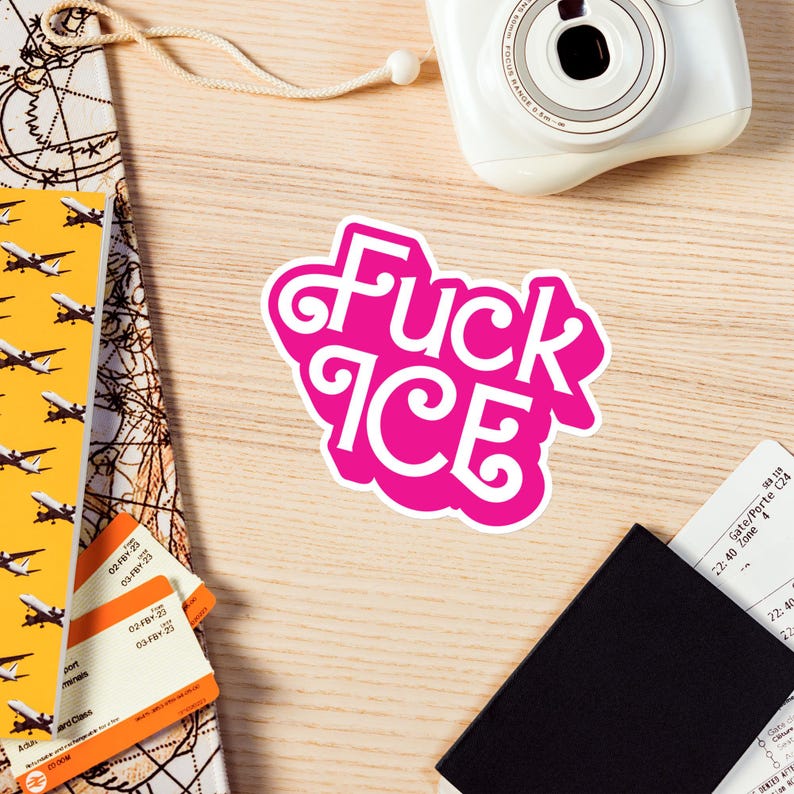 May include: A pink sticker with the words "Fuck ICE" in white, stylized lettering. The sticker is on a light wood surface, alongside a white camera, travel documents, and a passport.
