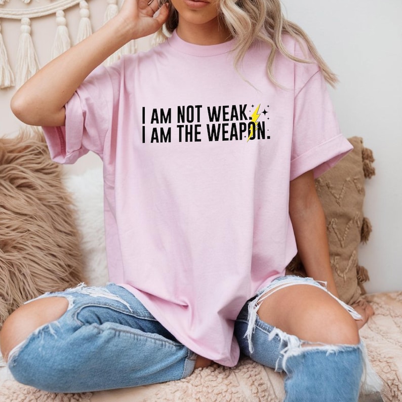 Puede incluir: Una camiseta rosa con el texto "I AM NOT WEAK. I AM THE WEAPON." en letras negras y un gr&aacute;fico de rayo amarillo.