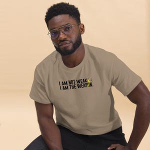 Puede incluir: Una persona vistiendo una camiseta marr&oacute;n claro con el texto "I AM NOT WEAK. I AM THE WEAPON." en letras negras y un s&iacute;mbolo de rayo amarillo.