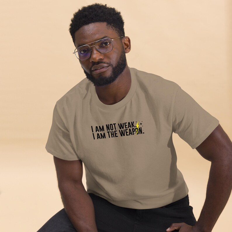 Puede incluir: Una persona vistiendo una camiseta marr&oacute;n claro con el texto "I AM NOT WEAK. I AM THE WEAPON." en letras negras y un s&iacute;mbolo de rayo amarillo.