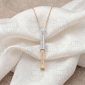 May include: A gold and silver bar pendant necklace with the text "YOUR Design Here" and "Hidden Message" engraved on it. The pendant hangs from a delicate gold chain, set against a white fabric background.