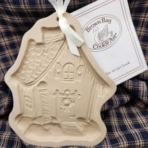 May include: A beige ceramic cookie mold shaped like a house with a textured roof, a door with a wreath, and a white ribbon tied through the top hole. Beside it is a white booklet with the text Brown Bag Cookie Art and a swan illustration.