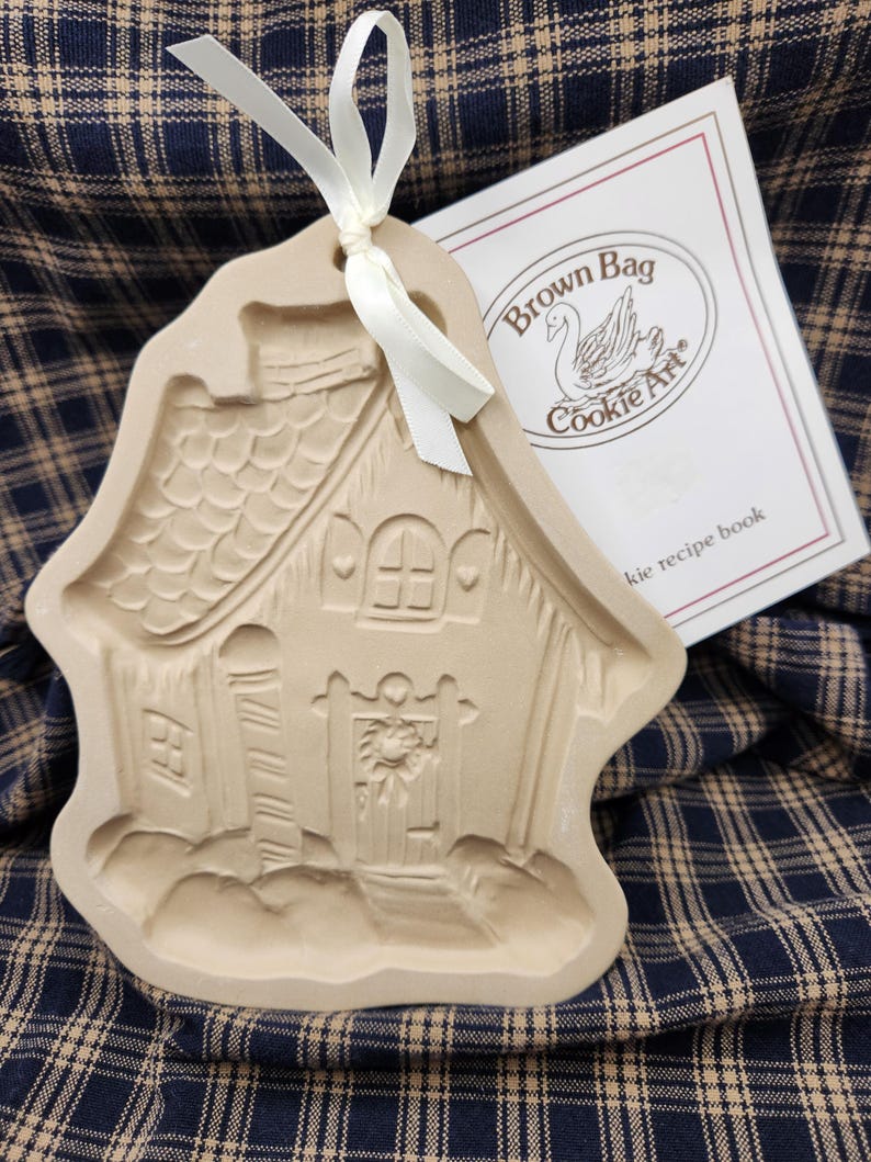 May include: A beige ceramic cookie mold shaped like a house with a textured roof, a door with a wreath, and a white ribbon tied through the top hole. Beside it is a white booklet with the text Brown Bag Cookie Art and a swan illustration.