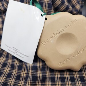 May include: A tan, scalloped-edge ceramic cookie mold with a central indentation. The text "BROWN BAG COOKIE ART" and "&copy;1988 HILL DESIGN" is embossed on the surface. A white paper tag is attached with a green ribbon, featuring printed text.