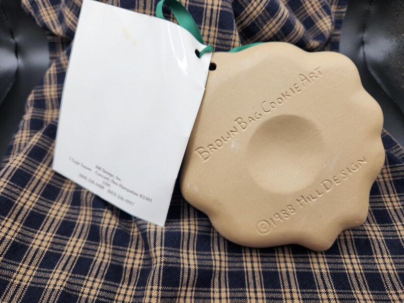 May include: A tan, scalloped-edge ceramic cookie mold with a central indentation. The text "BROWN BAG COOKIE ART" and "&copy;1988 HILL DESIGN" is embossed on the surface. A white paper tag is attached with a green ribbon, featuring printed text.