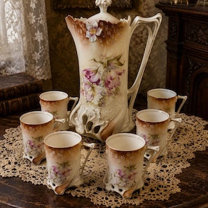 May include: A porcelain tea set consisting of a tall lidded pitcher and five matching mugs. Each piece features a cream-colored base with brown gradient rims, ornate sculpted bases, and hand-painted purple and white floral motifs with green leaves.