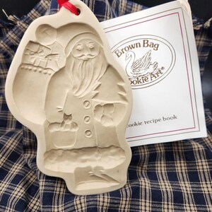 May include: A beige ceramic cookie mold featuring a relief of Santa Claus holding a small animal, suspended by a red ribbon. Beside it is a white booklet with the text Brown Bag Cookie Art and cookie recipe book printed on the front cover.