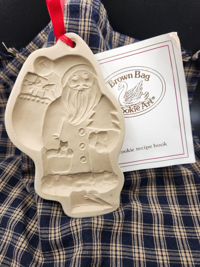 May include: A beige ceramic cookie mold featuring a relief of Santa Claus holding a small animal, suspended by a red ribbon. Beside it is a white booklet with the text Brown Bag Cookie Art and cookie recipe book printed on the front cover.