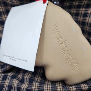 May include: A tan ceramic cookie mold with a smooth, matte finish. The surface is embossed with the text "BROWN BAG COOKIE ART &copy; 1989 HILL DESIGN". A white rectangular paper tag is attached to the top, featuring printed text and a red string.