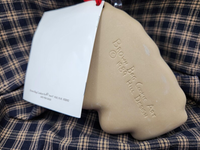 May include: A tan ceramic cookie mold with a smooth, matte finish. The surface is embossed with the text "BROWN BAG COOKIE ART &copy; 1989 HILL DESIGN". A white rectangular paper tag is attached to the top, featuring printed text and a red string.