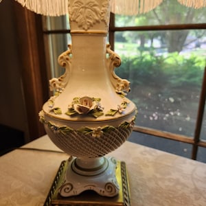 May include: A decorative table lamp with a cream-colored ceramic base and a fringed lampshade. The base features floral and leaf accents, with a gold-colored square base. The lamp's design includes a basket weave pattern and a rose.