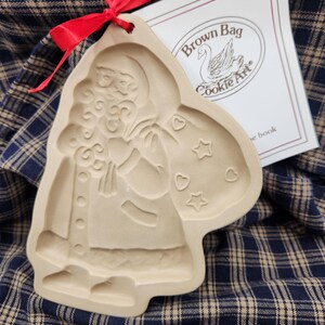 May include: A beige ceramic cookie mold featuring an embossed Santa Claus figure carrying a sack decorated with stars and hearts. A red ribbon is tied through a hole at the top. A white paper insert with the text Brown Bag Cookie Art is visible.