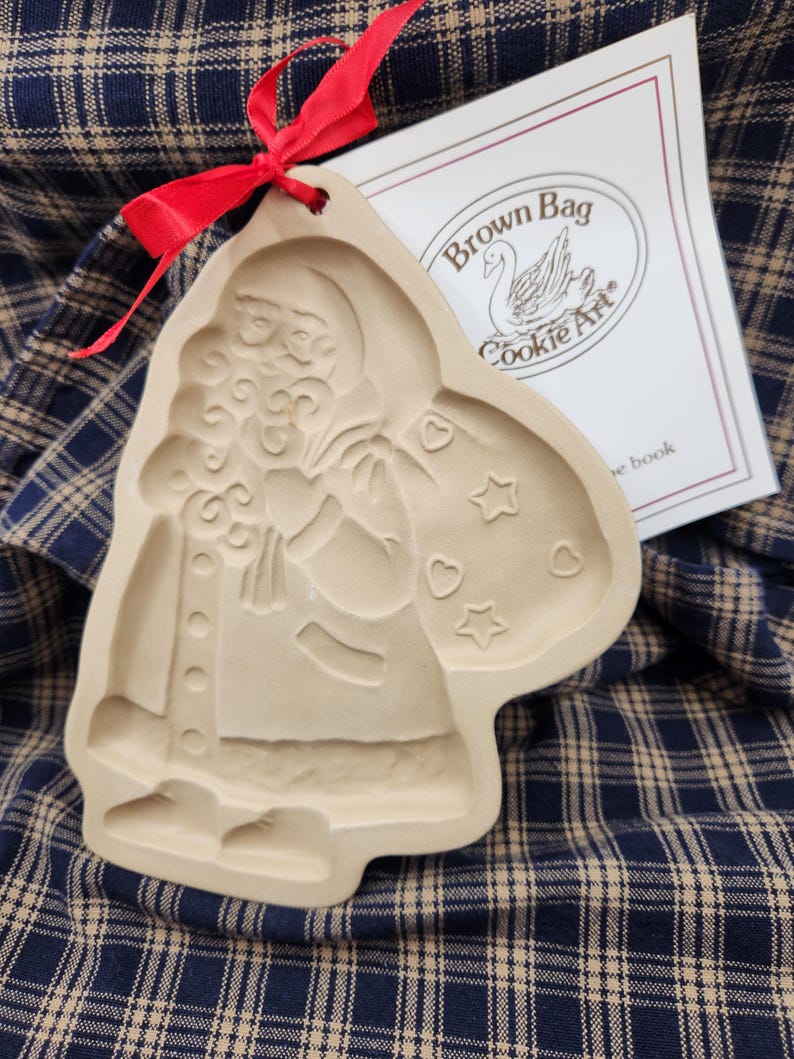 May include: A beige ceramic cookie mold featuring an embossed Santa Claus figure carrying a sack decorated with stars and hearts. A red ribbon is tied through a hole at the top. A white paper insert with the text Brown Bag Cookie Art is visible.
