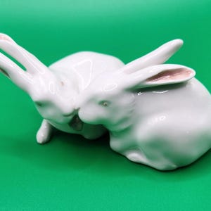 May include: A white ceramic figurine featuring two rabbits positioned side by side. The rabbits have smooth, glossy surfaces with delicate pink coloring on the inner parts of their long, upright ears. The sculpture captures the rabbits in a resting pose.