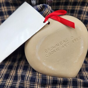 May include: A heart-shaped beige ceramic cookie mold with a red ribbon attached. The surface is embossed with the text BROWN BAG COOKIE ART 1985. A white rectangular paper tag is attached to the mold by the ribbon, featuring printed text.