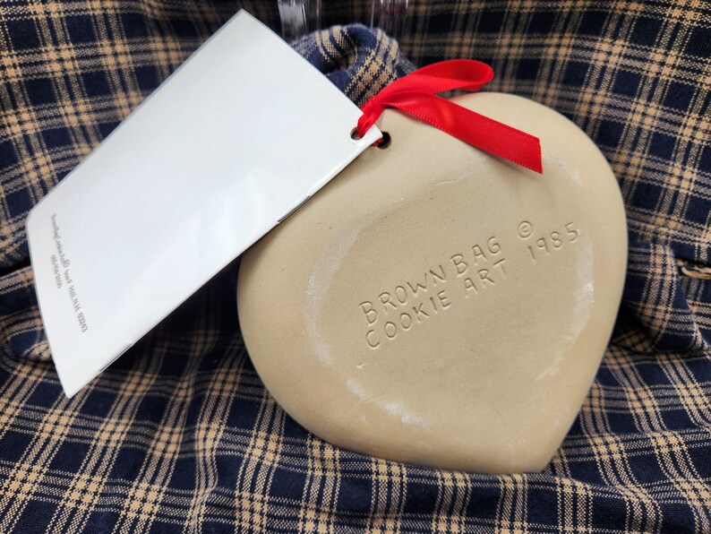 May include: A heart-shaped beige ceramic cookie mold with a red ribbon attached. The surface is embossed with the text BROWN BAG COOKIE ART 1985. A white rectangular paper tag is attached to the mold by the ribbon, featuring printed text.