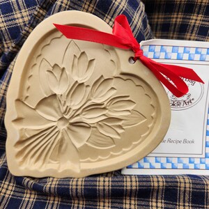 May include: A heart-shaped tan ceramic cookie mold featuring an embossed bouquet of tulips tied with a bow. A bright red ribbon is threaded through a hole at the top. Beside it is a white booklet with blue checkered borders and the text Cookie Art.