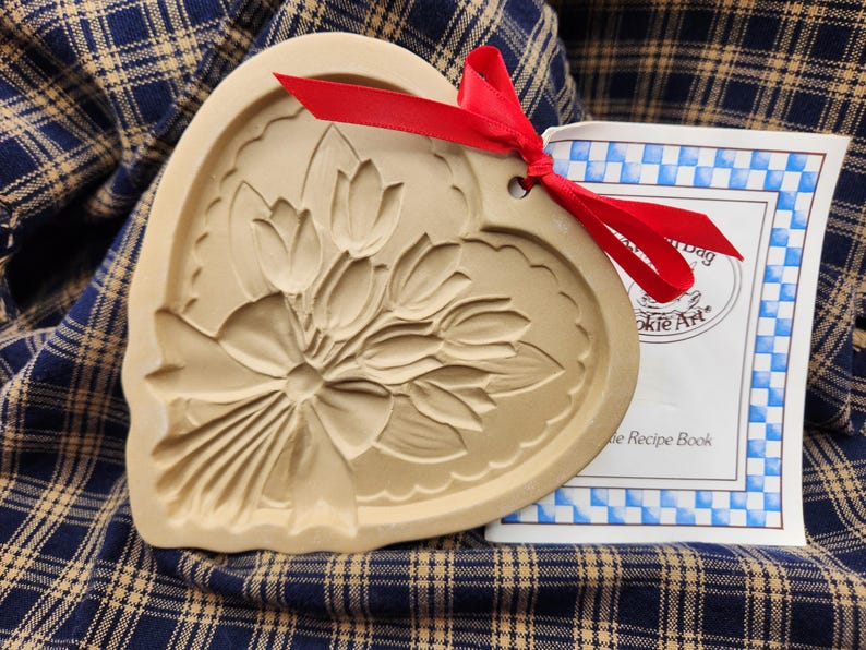 May include: A heart-shaped tan ceramic cookie mold featuring an embossed bouquet of tulips tied with a bow. A bright red ribbon is threaded through a hole at the top. Beside it is a white booklet with blue checkered borders and the text Cookie Art.