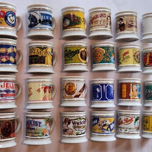 May include: A collection of twenty-four white ceramic mugs arranged in four rows of six. Each mug features a unique vintage-style advertisement or brand logo with colorful graphics and text, accented by a thin gold rim at the top and bottom edges.