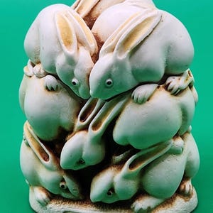 May include: A cream-colored sculpture featuring a cluster of five rabbits huddled together. The rabbits have smooth surfaces with subtle brown shading in the crevices. The base of the sculpture includes an oval plaque with the text P. CALVERT.