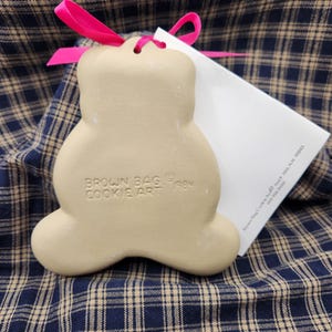 May include: A beige ceramic bear-shaped cookie mold with a hot pink ribbon tied through the top hole. The mold is embossed with the text BROWN BAG COOKIE ART 1984. A white informational card is tucked behind the mold, partially visible.