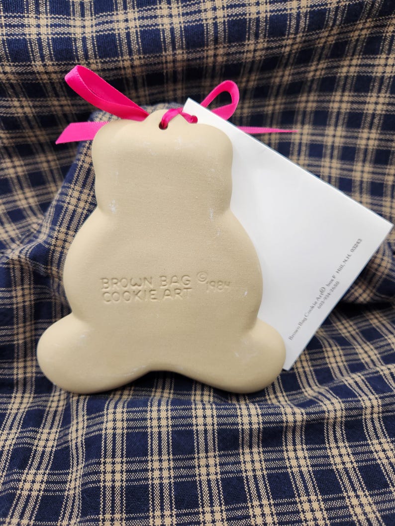 May include: A beige ceramic bear-shaped cookie mold with a hot pink ribbon tied through the top hole. The mold is embossed with the text BROWN BAG COOKIE ART 1984. A white informational card is tucked behind the mold, partially visible.