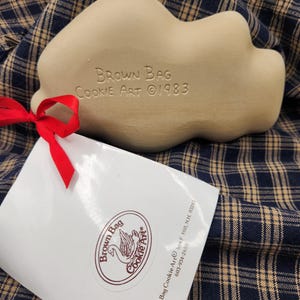 May include: A beige ceramic cookie mold with the embossed text Brown Bag Cookie Art 1983. A red ribbon is tied to the mold, attaching a white paper tag featuring the Brown Bag Cookie Art logo, address, and phone number in dark brown ink.