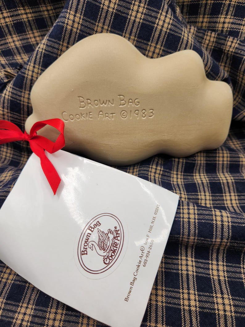 May include: A beige ceramic cookie mold with the embossed text Brown Bag Cookie Art 1983. A red ribbon is tied to the mold, attaching a white paper tag featuring the Brown Bag Cookie Art logo, address, and phone number in dark brown ink.