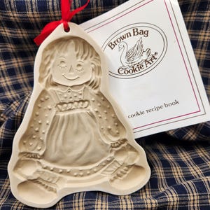 May include: A beige ceramic cookie mold shaped like a doll with a red ribbon tied through a hole at the top. Next to it is a white booklet with the text Brown Bag Cookie Art and a swan illustration, along with the words cookie recipe book.