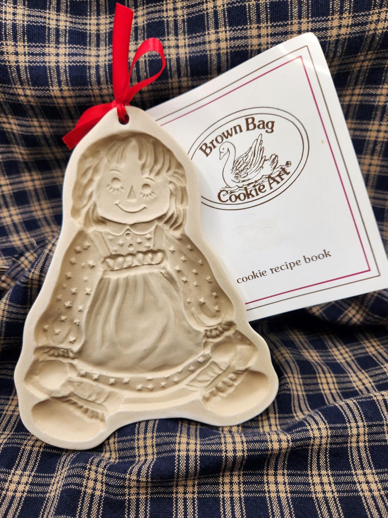 May include: A beige ceramic cookie mold shaped like a doll with a red ribbon tied through a hole at the top. Next to it is a white booklet with the text Brown Bag Cookie Art and a swan illustration, along with the words cookie recipe book.