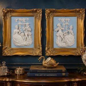 May include: Two rectangular wall plaques featuring white relief figures of people playing musical instruments against a light blue background, encased in ornate gold frames. The plaques are displayed above a surface with books and decor.