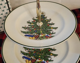Puede incluir: Un soporte de dos niveles de porcelana blanca con borde verde y un asa central de metal plateado. Cada plato muestra una ilustración colorida de un árbol de Navidad decorado rodeado de varios regalos envueltos y un coche de juguete azul.