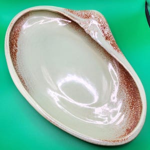 May include: An oval ceramic serving dish with a smooth, light cream-colored center and a textured, speckled reddish-brown rim. The dish features a slightly raised, asymmetrical edge on one side, creating a distinct, organic, and elongated shape.