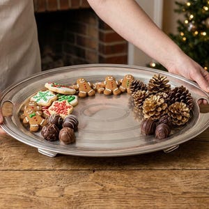 May include: A large, oval, silver-toned metal serving tray with integrated side handles and a raised rim. The tray holds a collection of gingerbread men, snowflake-shaped cookies, round chocolate truffles, and several natural pinecones.