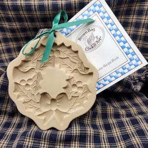 May include: A beige ceramic cookie mold featuring an embossed wreath design with holly leaves, berries, and a central bow, accented by a green ribbon tied through the top hole, accompanied by a white booklet labeled Brown Bag Cookie Art.