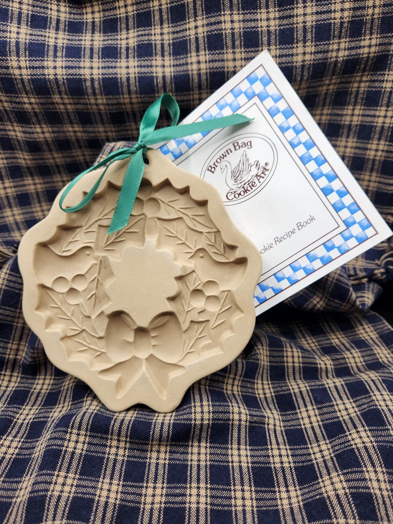 May include: A beige ceramic cookie mold featuring an embossed wreath design with holly leaves, berries, and a central bow, accented by a green ribbon tied through the top hole, accompanied by a white booklet labeled Brown Bag Cookie Art.