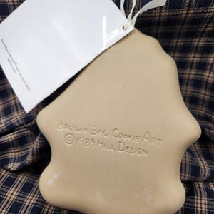 May include: A tan ceramic cookie mold with a smooth, rounded shape. The surface is embossed with the text "BROWN BAG COOKIE ART &copy; 1989 HILL DESIGN". A white paper tag is attached to the top with a thin white ribbon, and the mold has a flat back.