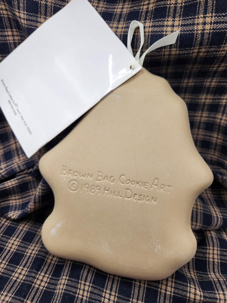 May include: A tan ceramic cookie mold with a smooth, rounded shape. The surface is embossed with the text "BROWN BAG COOKIE ART &copy; 1989 HILL DESIGN". A white paper tag is attached to the top with a thin white ribbon, and the mold has a flat back.