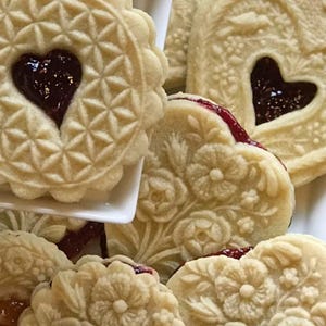 May include: A collection of round and heart-shaped shortbread cookies featuring intricate embossed floral and geometric patterns. Each cookie has a central cutout filled with dark red fruit jam, revealing a deep crimson center.