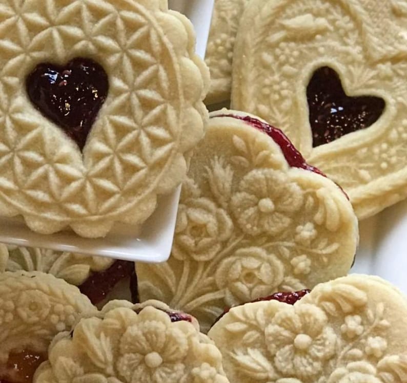 May include: A collection of round and heart-shaped shortbread cookies featuring intricate embossed floral and geometric patterns. Each cookie has a central cutout filled with dark red fruit jam, revealing a deep crimson center.
