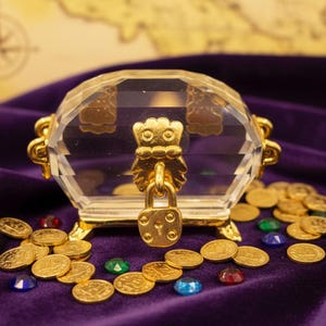 May include: A clear, faceted treasure chest with gold accents and a padlock, resting on a purple velvet surface. Gold coins and colorful gems surround the chest, with a map in the background. A decorative item.