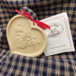 May include: A heart-shaped beige ceramic cookie mold featuring an embossed floral design with tulips and small hearts, tied with a red ribbon. Beside it is a white booklet with the text Brown Bag Cookie Art and cookie recipe book.