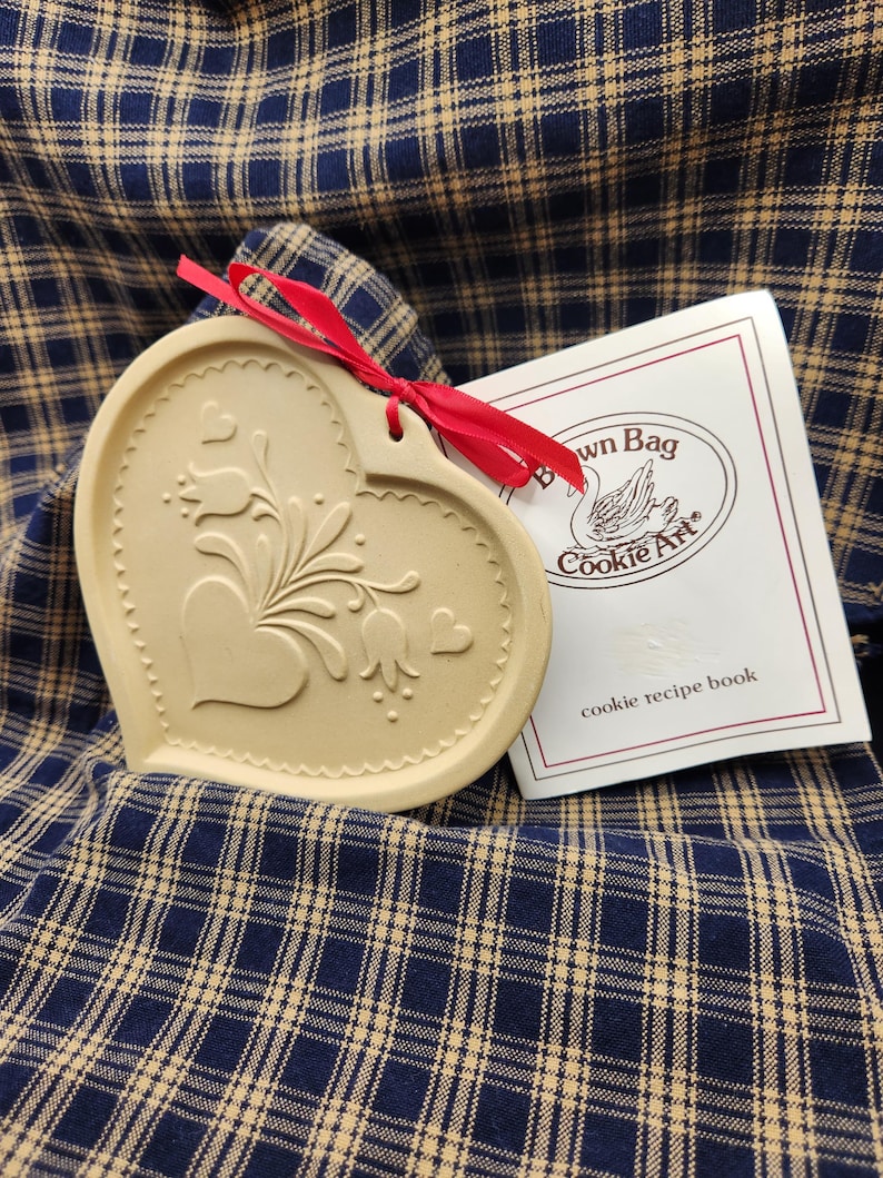 May include: A heart-shaped beige ceramic cookie mold featuring an embossed floral design with tulips and small hearts, tied with a red ribbon. Beside it is a white booklet with the text Brown Bag Cookie Art and cookie recipe book.