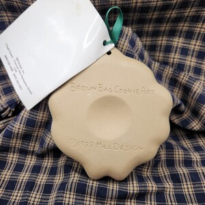 May include: A beige ceramic cookie mold with a scalloped edge and a green ribbon loop. The text "BROWN BAG COOKIE ART" and "&copy;1988 HILL DESIGN" is embossed on the surface. A white paper tag is attached to the mold with information about Hill Design.