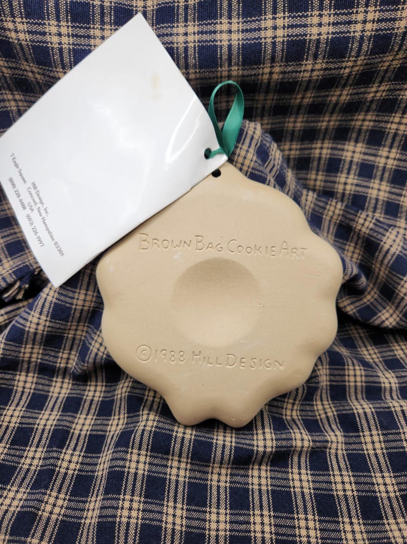 May include: A beige ceramic cookie mold with a scalloped edge and a green ribbon loop. The text "BROWN BAG COOKIE ART" and "&copy;1988 HILL DESIGN" is embossed on the surface. A white paper tag is attached to the mold with information about Hill Design.