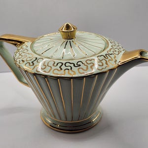 May include: A light green teapot with a fluted body and gold trim. The lid features a gold finial and a gold scroll pattern. The handle and spout are accented with gold, and the base is circular with a gold rim, creating a cohesive design.