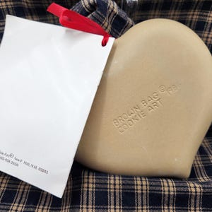 May include: A tan heart-shaped ceramic cookie mold with the embossed text BROWN BAG COOKIE ART 1986. A white rectangular tag with black text and a red ribbon is attached to the mold. The tag includes the text Brown Bag Cookie Art, box F, Hill, N.H. 03243, 603-934-2650.