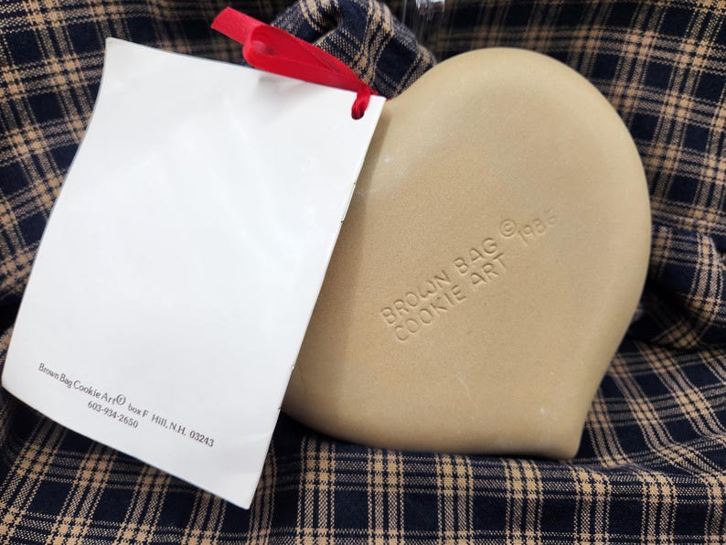 May include: A tan heart-shaped ceramic cookie mold with the embossed text BROWN BAG COOKIE ART 1986. A white rectangular tag with black text and a red ribbon is attached to the mold. The tag includes the text Brown Bag Cookie Art, box F, Hill, N.H. 03243, 603-934-2650.