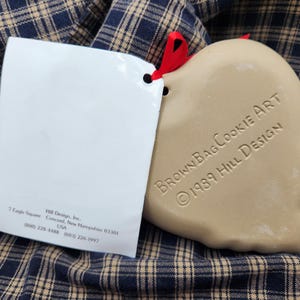 May include: A tan heart-shaped ceramic cookie mold with a red ribbon, featuring the embossed text BrownBagCookieArt 1989 Hill Design. Attached is a white paper tag with the address 7 Eagle Square, Concord, New Hampshire 03301 and phone numbers.